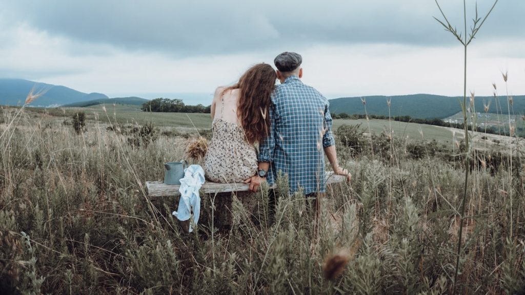 Portrait photo session of bride and groom on the field of village in country style. Light tone wedding on nature among dry grass. Man and woman are wear country clothes sitting on bench cute talking.